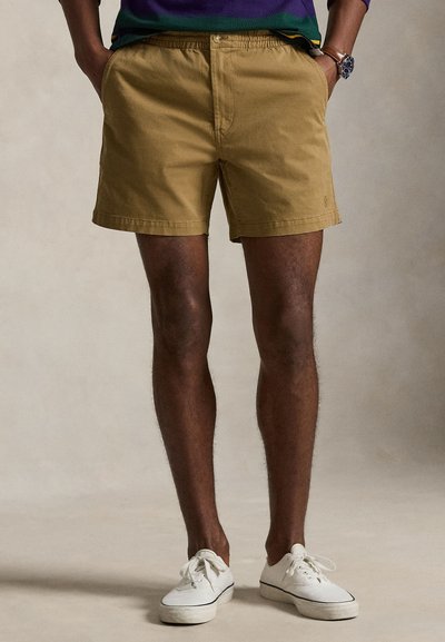 Uomo che indossa pantaloncini beige, scarpe da ginnastica bianche e una maglietta a righe viola, verde e blu navy, con le mani nelle tasche.