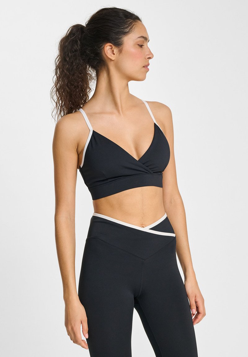 Svart sport-bh med v-formad design, kontrasterande vita band, tillverkad av mjukt tyg. Bärs med svarta högmidjade leggings.