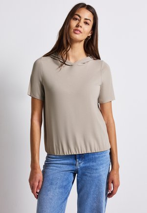 Haut beige à manches courtes avec capuche, offrant une coupe décontractée et un ourlet élastique. Associé à un jean bleu clair. Texture de tissu lisse.