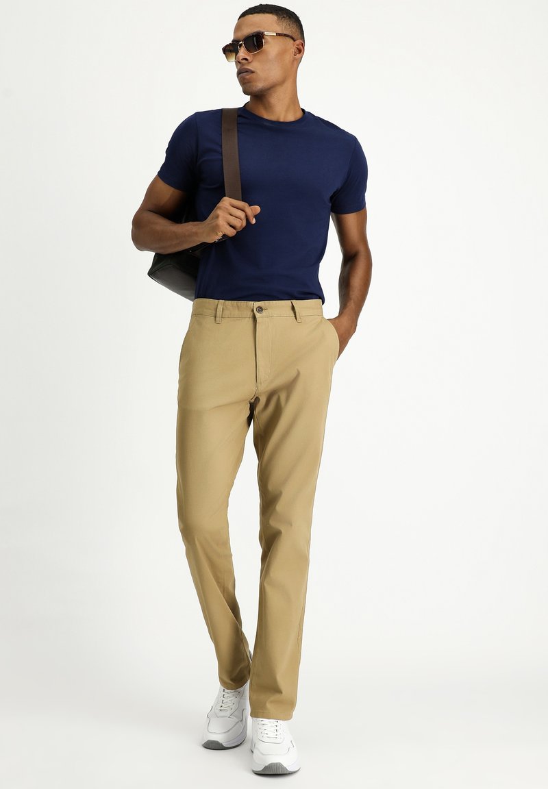 T-shirt attillata blu navy abbinata a pantaloni chinos beige e sneakers bianche. L'uomo porta una borsa nera, indossando occhiali da sole, con un design pulito e minimal.