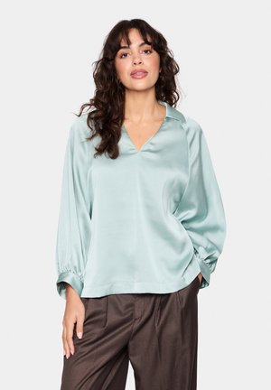 Light mint green satin blouse with a V-neck and puffed long sleeves, featuring a relaxed fit and gathered cuffs. Worn with brown trousers.