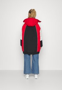 Person med blond hestehale iført en oversized rød, sort og hvid hættetrøje, brede blå jeans og hvide sneakers, som står mod en hvid baggrund.