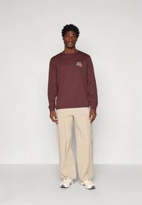 Bordeauxrode T-shirt met lange mouwen en logodetail op de linkerborst, gecombineerd met beige loszittende broek en lichtgekleurde sneakers.