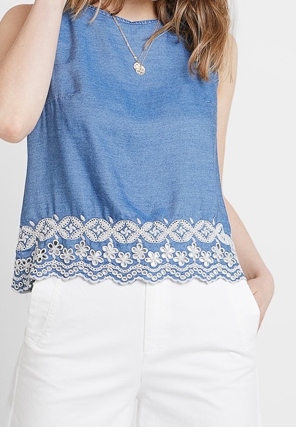 Femme portant un haut sans manches bleu avec une bordure en dentelle florale blanche à l'ourlet et un pantalon blanc, une main dans la poche.