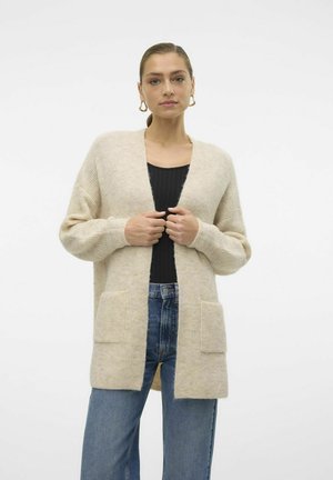 Beige gestrickte Strickjacke mit langen Ärmeln, offenem Vorderteil und zwei Vorderaschen, kombiniert mit einem schwarzen Oberteil und blauen hoch geschnittenen Jeans.