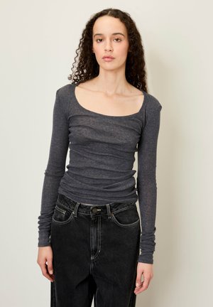 Haut à manches longues, de couleur gris foncé, avec un décolleté en ronde et une texture froncée. Associé à un jean noir taille haute avec poches avant.