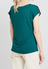 Mujer con una blusa de manga corta color aguamarina con acentos negros en los hombros y pantalones color crema, vista desde atrás contra un fondo liso.
