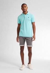 Lichtblauw poloshirt met korte mouwen, gecombineerd met grijze shorts met opgerolde zoom. Model draagt witte sneakers en staat op een neutrale achtergrond.