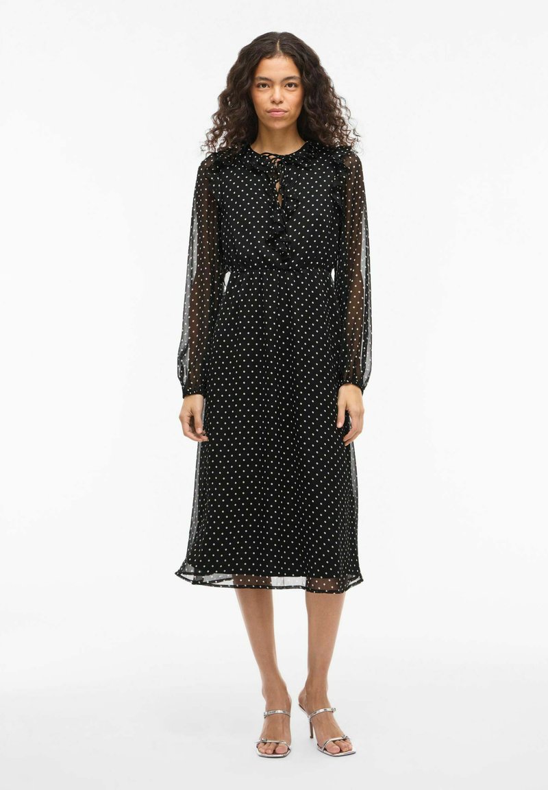 Robe noire à pois blancs avec des manches longues transparentes, un col froncé et une taille cintrée. Le tissu est léger et arrive au niveau des mollets.