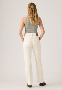 Γυναίκα με μακριά ξανθά μαλλιά, φοράει γκρι αμάνικη μπλούζα, ψηλόμεσα κρεμ τζιν και σκούρα καφέ παπούτσια, στέκεται μπροστά σε απλό φόντο.