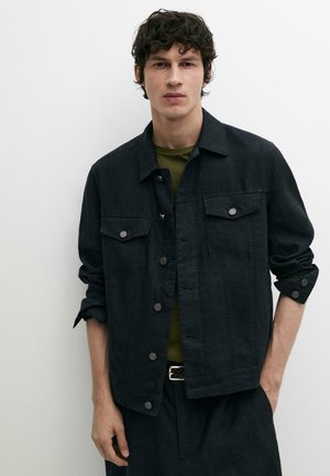 Junger Mann mit lockigem dunklem Haar, der eine schwarze, geknöpfte Jacke über einem olivgrünen Hemd trägt und mit einer Hand in der Tasche steht.