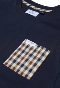 T-shirt blu navy con una tasca applicata a quadri che presenta quadratini marroni, beige e blu. Materiale in cotone morbido con scollatura rotonda.