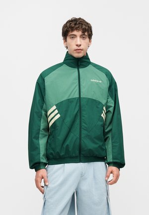 Ung mand iført en grøn Adidas trackjakke med hvide striber og lyseblå cargobukser, stående mod en ensfarvet hvid baggrund.