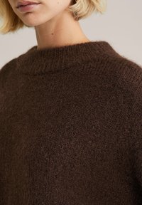 Pulover tricotat maro, cu un guler înalt, cu nervuri și o textură moale, pufoasă. Materialul prezintă un ușor luciu și o textură groasă.