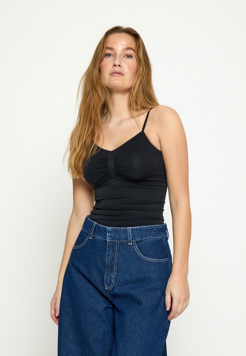 Jonge vrouw met lang lichtbruin haar, gekleed in een zwart gerimpeld spaghetti-strap topje en losse blauwe spijkerbroek, staand tegen een effen witte achtergrond.