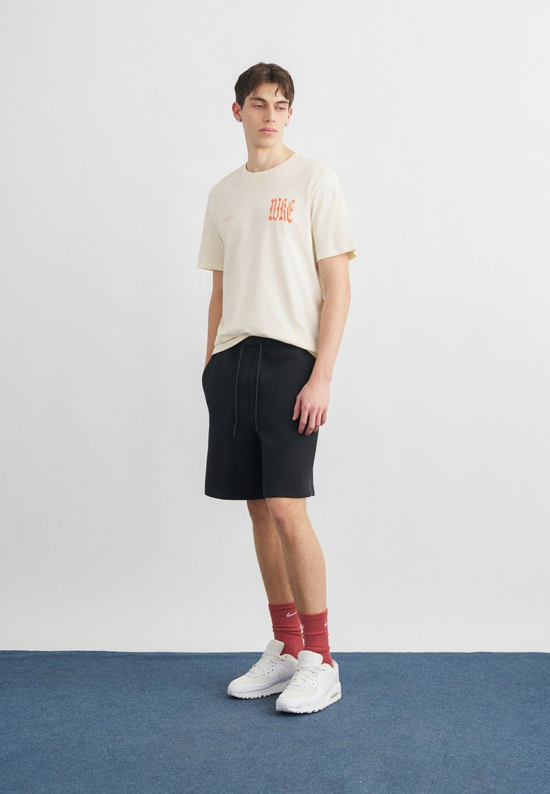 Jeune homme portant un t-shirt graphique beige, un short noir à cordon, des chaussettes rouges montantes et des baskets blanches, debout sur un tapis bleu.