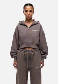 Bruine cropped sweatshirt met een capuchon en een ritssluiting aan de voorkant, met de tekst "GOODFORNOTHING" in het wit, gecombineerd met bijpassende sportbroeken.
