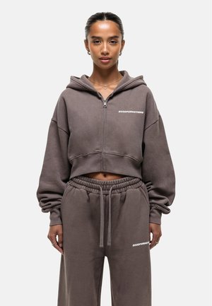 Bruine cropped sweatshirt met een capuchon en een ritssluiting aan de voorkant, met de tekst "GOODFORNOTHING" in het wit, gecombineerd met bijpassende sportbroeken.