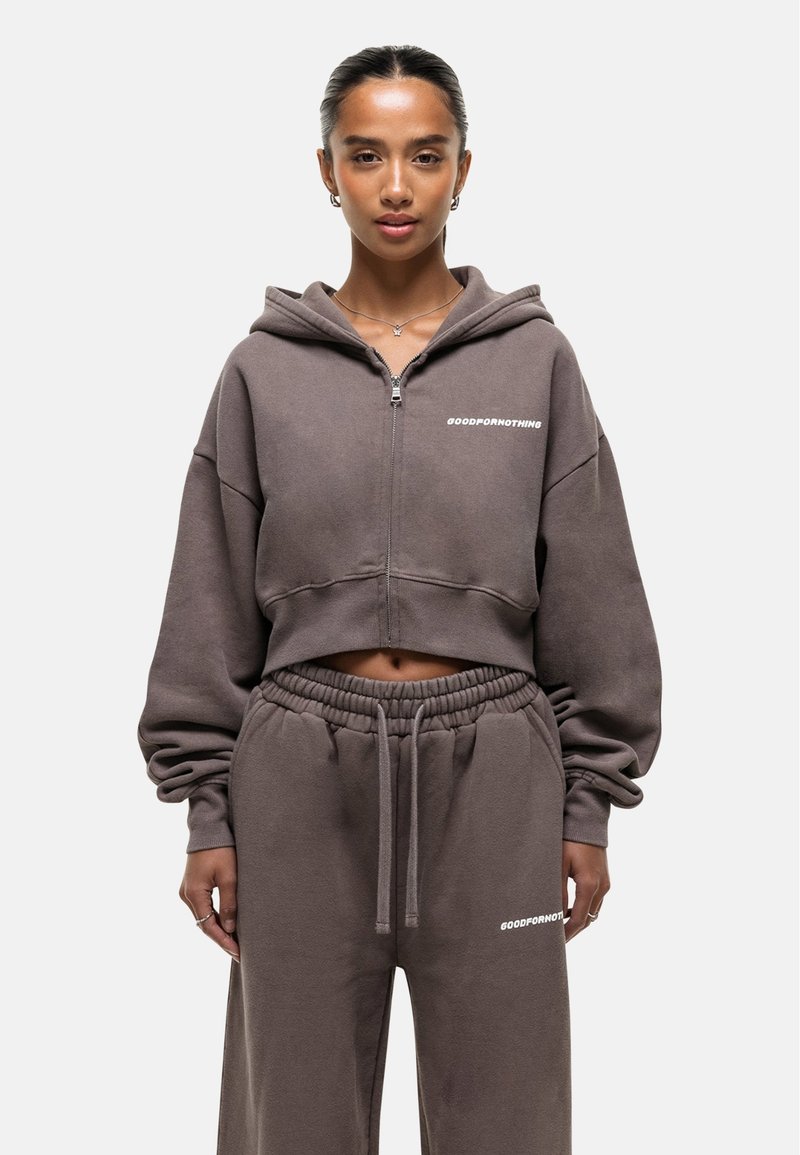 Bruine cropped sweatshirt met een capuchon en een ritssluiting aan de voorkant, met de tekst "GOODFORNOTHING" in het wit, gecombineerd met bijpassende sportbroeken.