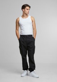 Black cargo pants with two side pockets, paired with a white tank top and white sneakers. The fabric appears sturdy with a relaxed fit.