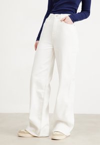 Jeans blancos de pierna ancha con una cintura alta, diseño de cinco bolsillos y una textura suave, combinados con zapatillas en tonos beige y crema.