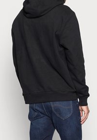 Sudadera negra de tejido suave con un ajuste relajado, que presenta una capucha de doble capa, puños acanalados y cintura ajustada. Combinada con jeans de color azul oscuro.