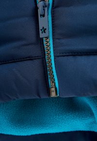 Veste matelassée bleu marine avec une fermeture éclair turquoise contrastante. Dotée d'une fermeture éclair en plastique proéminente avec un tirette en forme d'étoile et d'une doublure en polaire.