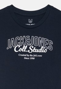 Tamsiai mėlynas medvilninis marškinėlis su apvaliu iškirpimu ir ryškia baltu tekstu: "JACK & JONES Coll. Studio" bei papildomu prekių ženklu žemiau.