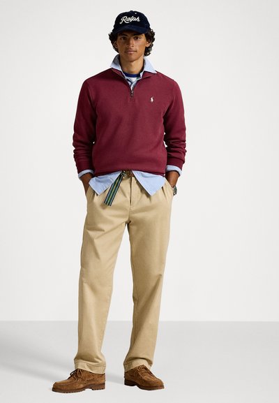 Tânăr îmbrăcat cu un pulover burgund cu fermoar la jumătate, cămașă bleu deschis, pantaloni bej cu curea cu dungi, pantofi maro și o șapcă bleumarin cu "Ralph".