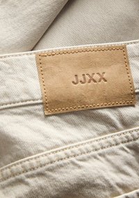 Tissu beige à texture lisse avec un tissage visible, doté d'une étiquette en cuir marron embossée du texte "JJXX." Des détails cousus viennent accentuer le tissu.