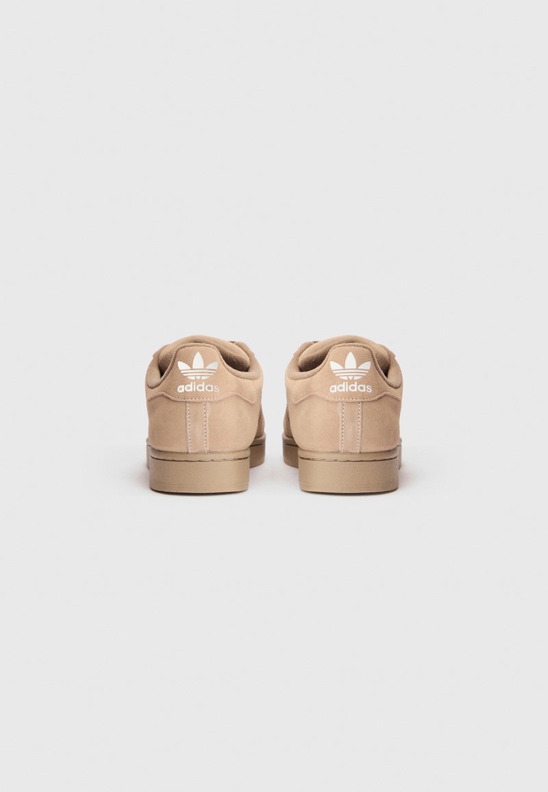 Baskets en suède brun avec un design basse, arborant un logo blanc au niveau du talon. Semelle en caoutchouc clair et détails de couture contrastants.
