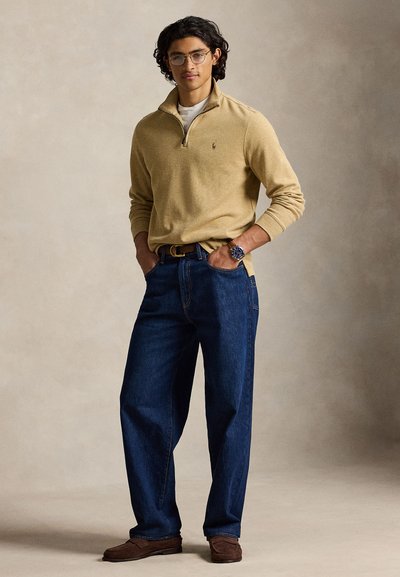 Jeune homme portant un pull beige à fermeture éclair quart de longueur, un jean bleu, des chaussures marron, des lunettes et une montre-bracelet, debout les mains dans les poches.