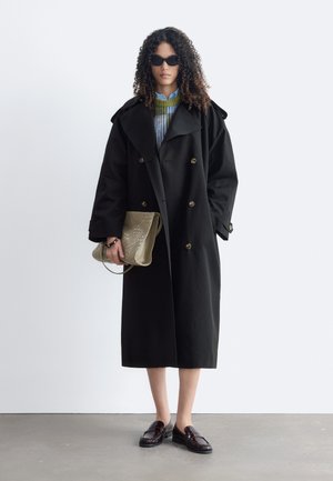 Femme aux cheveux bouclés portant des lunettes de soleil, un long manteau noir, un pull à rayures, tenant une pochette beige texturée et des mocassins foncés.