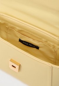 Sac à main jaune ouvert pour montrer une poche intérieure en tissu et une étiquette noire avec le nom de la marque "ANNA FIELD" à l'intérieur.