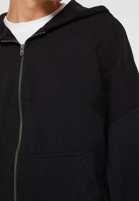 Sudadera negra con cremallera fabricada en una mezcla de algodón. Presenta una capucha, bolsillo frontal y detalle de cremallera metálica visible. Diseño minimalista.