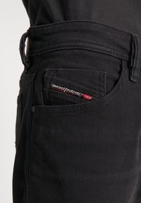 Black denim jeans close-up showing the front pocket with a small Diesel Industry label in black, white, and red stitching.