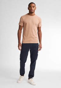 Krátký rukáv hnědého trička v kombinaci s tmavě modrými cargo kalhotami s bočními kapsami, doplněné krémovými teniskami. Jednoduchý, neformální outfit.