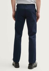 Chino bleu marine avec une coupe droite, deux poches arrière et une texture de tissu. Porté avec des chaussures noires, pris de l'arrière.
