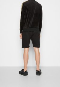 Svart velourjacka med guldlinjer på sidorna, kombinerad med svarta shorts. Jackan har en hög krage och elastiska muddlar. Svarta sneakers fulländar looken.