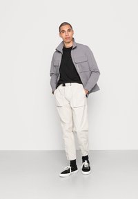 Giacca zip grigia chiara con due tasche frontali, T-shirt nera, pantaloni cargo crema e sneaker nere con suola bianca.