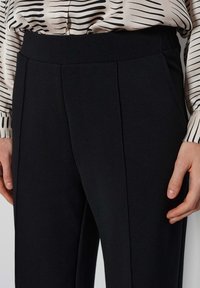 Pantalon noir ajusté avec des plis sur le devant, une ceinture élastique et une texture lisse. Porté avec un chemisier à motifs présentant des rayures.