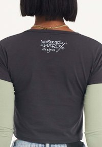 Dunkelgraues, kurzärmeliges Cropped-T-Shirt mit weißem Aufdruck des Logos "DON ED HARDY designs" auf dem oberen Rücken. Lange grüne Ärmel darunter.