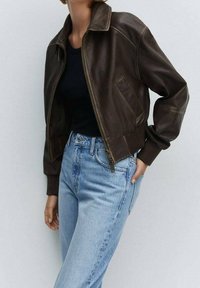 Femme portant une veste aviateur en cuir marron foncé sur un haut noir et un jean taille haute bleu clair, debout devant un fond uni.