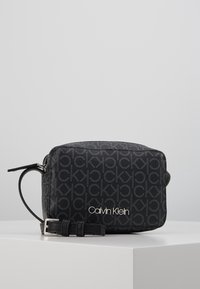 Černá obdélníková crossbody taška s tištěným vzorem loga, hladkým povrchem a stříbrným prvkem názvu značky na přední straně.