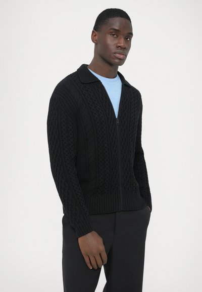 Pull noir en maille tricotée avec fermeture éclair, présentant des motifs en torsades texturées, un col à revers, des poignets et un ourlet côtelés, porté sur une chemise bleue.