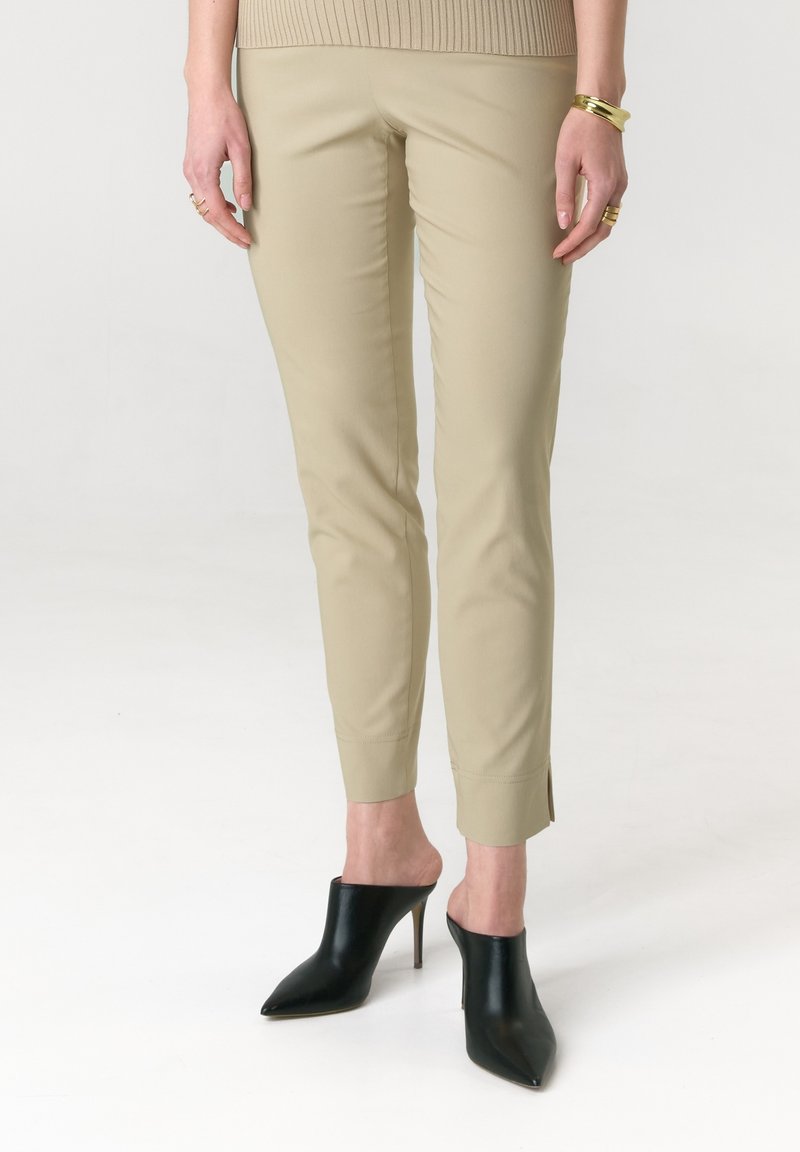 Beige hautgeschnittene Hosen mit glatter Textur, knöchellang und seitlichen Schlitzen. Kombiniert mit eleganten schwarzen Mules mit spitzen Zehen und hohen Absätzen.