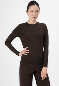 Tânără cu păr închis la culoare, îmbrăcată cu un top lung cu mâneci maro și pantaloni întunecați cu un model, stând cu o mână pe șold, în fața unui fundal alb.