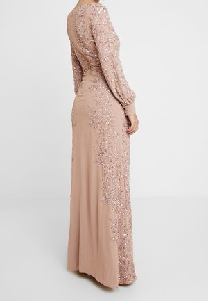 Femme portant une longue robe rose blush avec broderies florales en sequins, manches longues et dos plongeant, tournée de dos sur un fond uni.