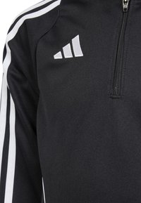 Melnā sportiskā jaka ar rāvējslēdzēju, ar baltu trīsrindu dizainu, teksturētu audumu un logo uzlīmi augšējā kreisajā pusē.