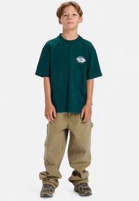 Roheline puuvillane T-särk lühikeste varrukatega ja ümar kaelusega, mille rinnal on valge graafiline trükk. Sobitatud beežide lahtiste taskupükstega ja mustriliste jalanõudega.